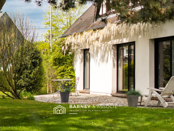 White-walled house with a cobblestone patio, cascading white blossoms along the roof, and a small table with chairs in a sunny garden.