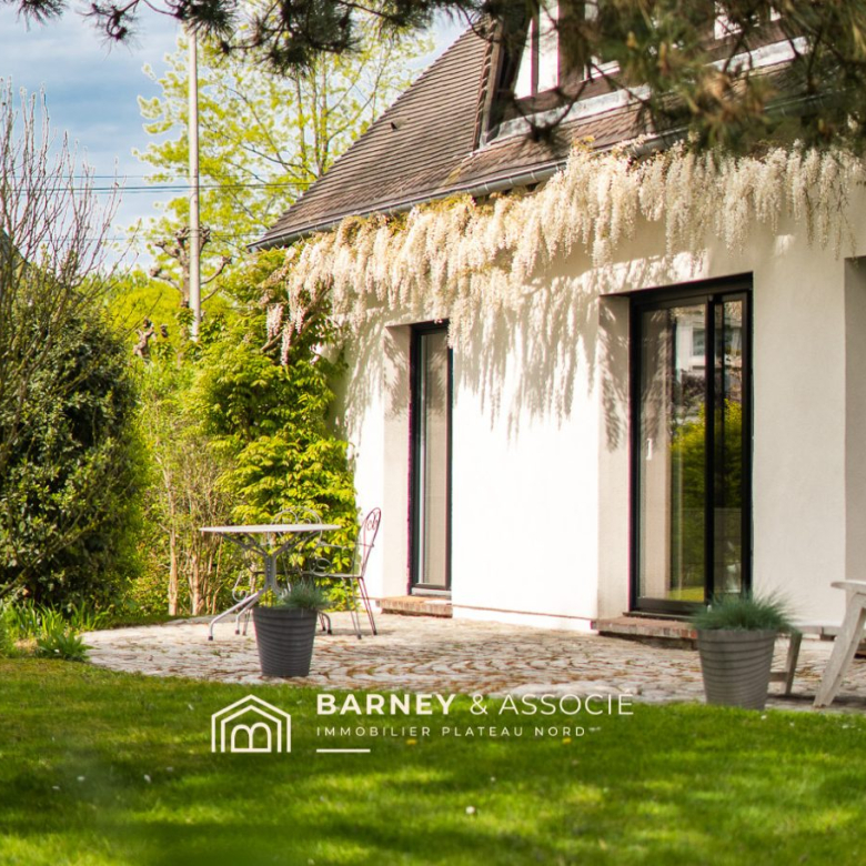 White-walled house with a cobblestone patio, cascading white blossoms along the roof, and a small table with chairs in a sunny garden.