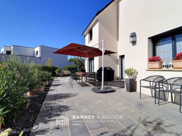 Terrace of a contemporary house with garden furniture and parasol, surrounded by greenery.