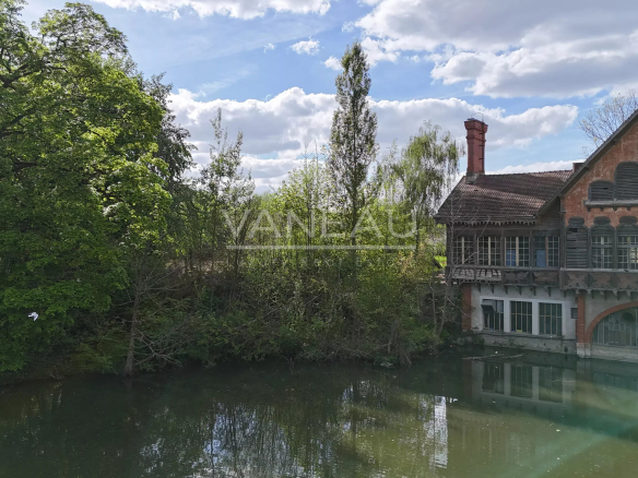 Old brick house with arched windows by a calm river, surrounded by green trees under a blue, partly cloudy sky.