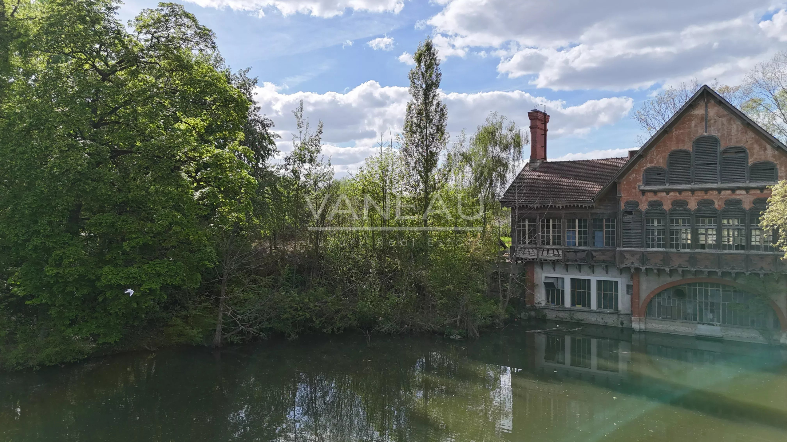 447a5fd04e97f74a2b44c7cb3c3aa3f2_35b1e3af66_4032-original Old brick house with arched windows by a calm river, surrounded by green trees under a blue, partly cloudy sky.