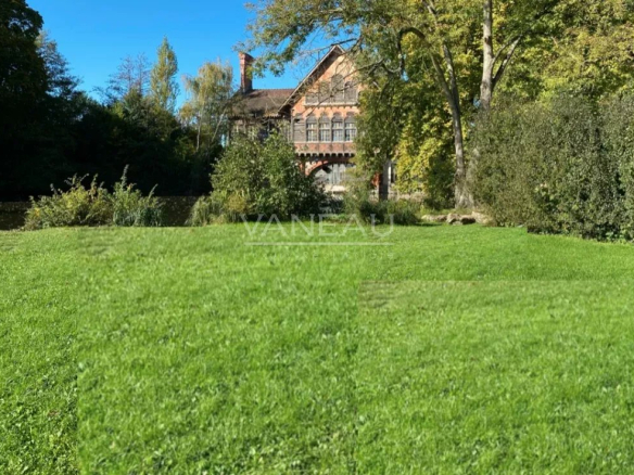 Brick house with a gabled roof behind a lush green lawn and trees on a sunny day.
