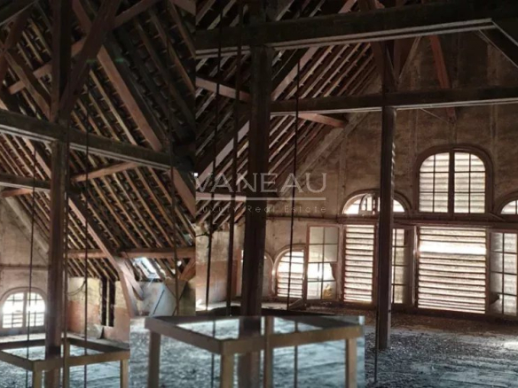 Attic-like interior with exposed wooden roof trusses, dark columns, and large arched windows; two white display tables in front.