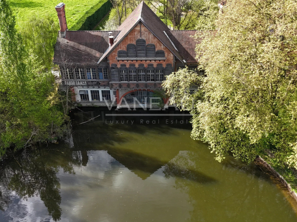 Aerial view of an old brick house by a calm river, surrounded by dense green trees and reflecting on the water surface.