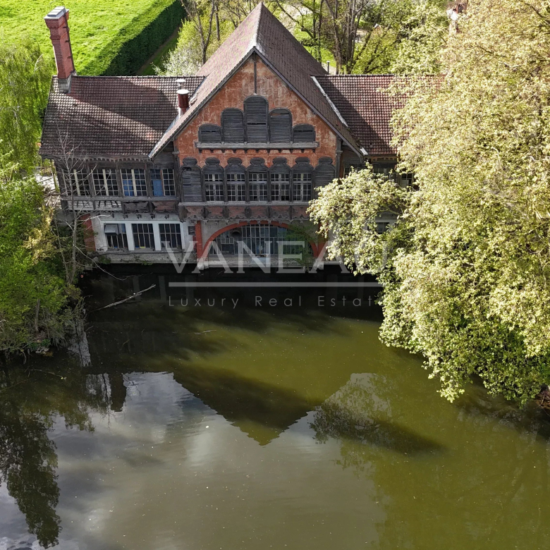 Aerial view of an old brick house by a calm river, surrounded by dense green trees and reflecting on the water surface.