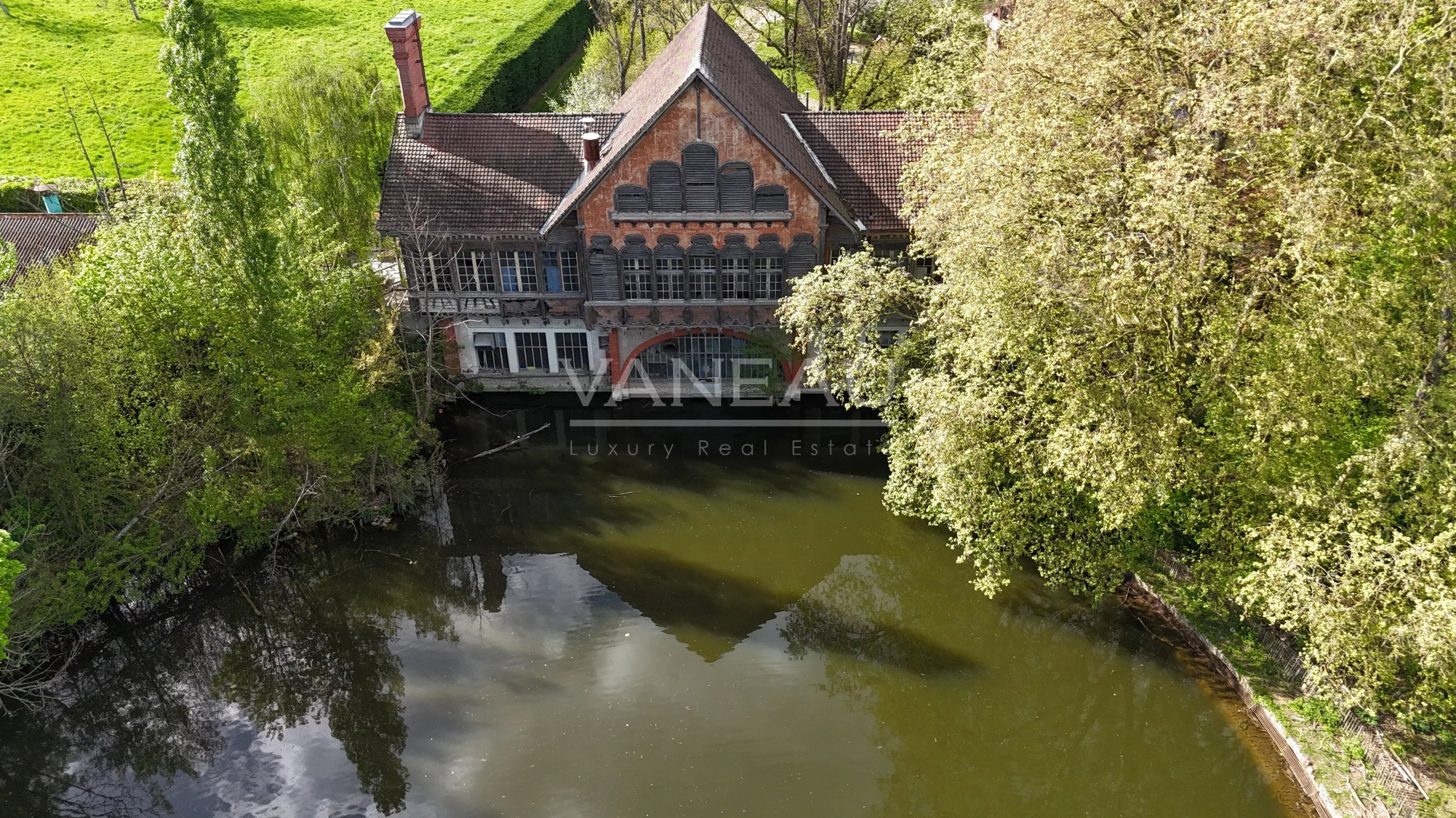 Aerial view of an old brick house by a calm river, surrounded by dense green trees and reflecting on the water surface.