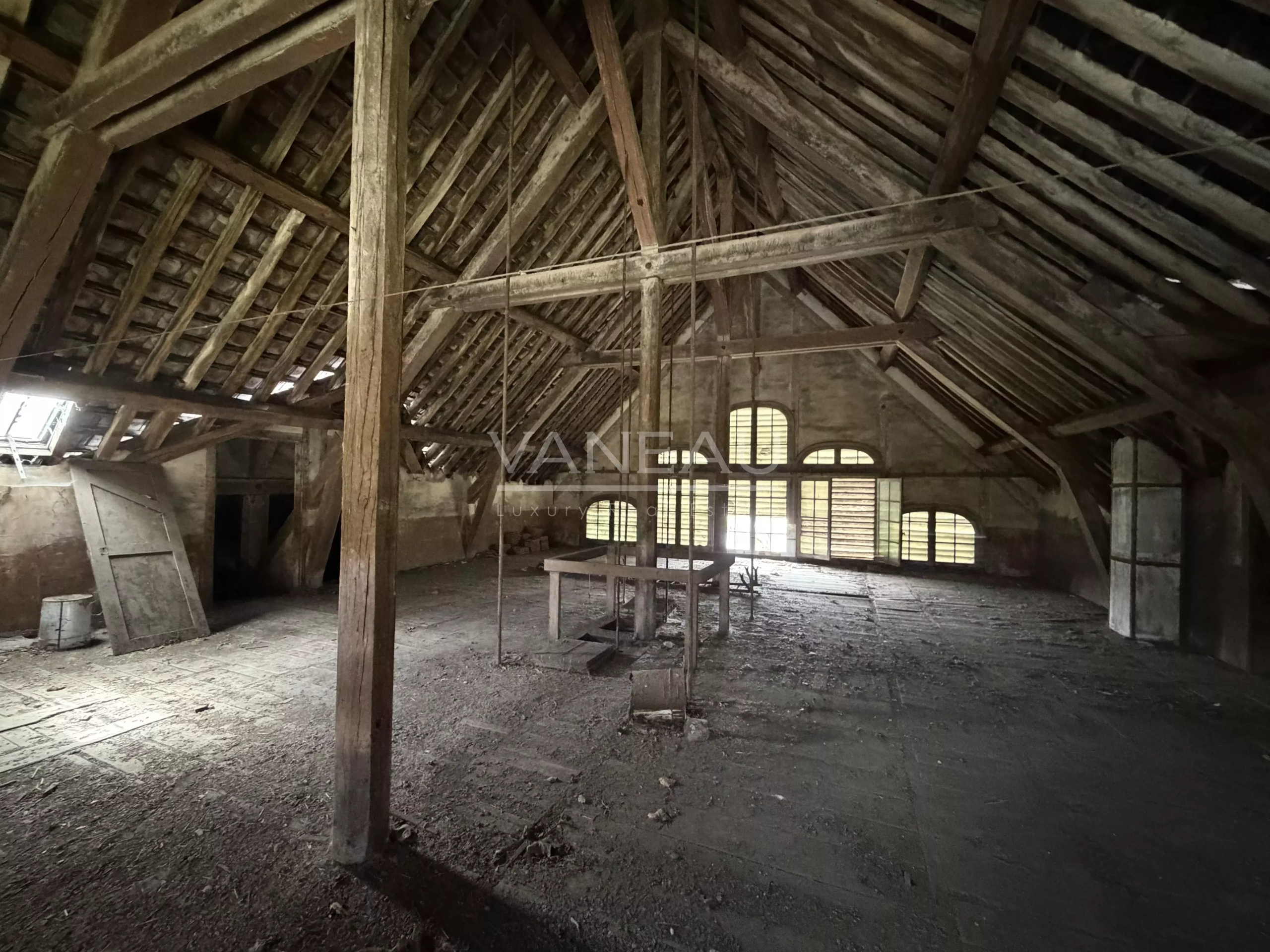90ade101ac9128f844aeb71f0790f315_27ed5499bc_4096-original Dusty attic with exposed wooden beams and a central worktable under a high, sloped ceiling; light filters through arched windows at the far end.