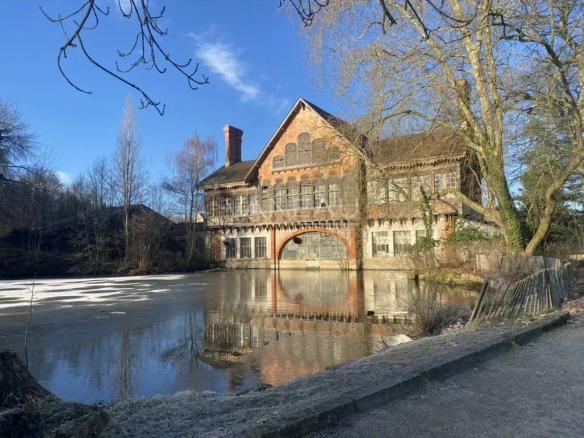 Brick Victorian-era mill by a reflective, partially frozen pond; bare trees frame the brick building under a blue sky