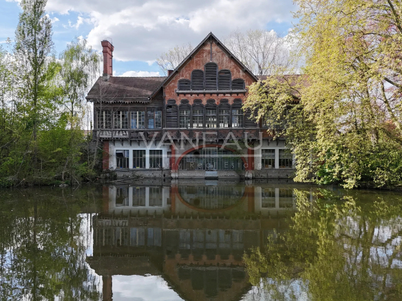 Red brick mansion with a steep gabled roof and arched windows beside a calm lake, reflected in the water, surrounded by trees.