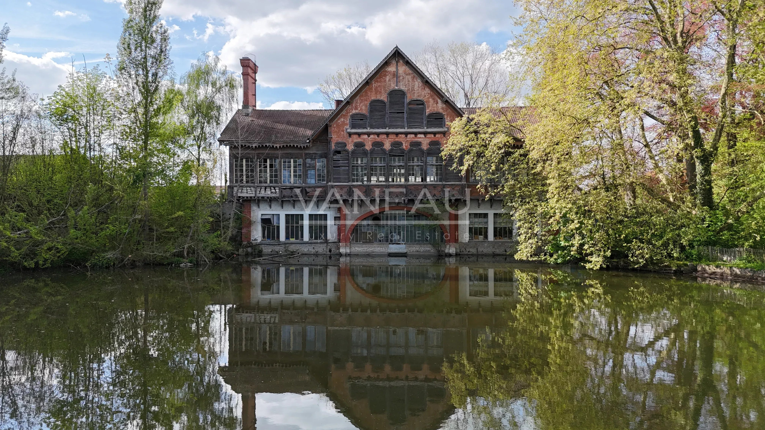 Red brick mansion with a steep gabled roof and arched windows beside a calm lake, reflected in the water, surrounded by trees.