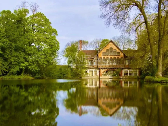 Brick riverside house with a gabled roof, nestled among green trees and reflected in a calm lake.