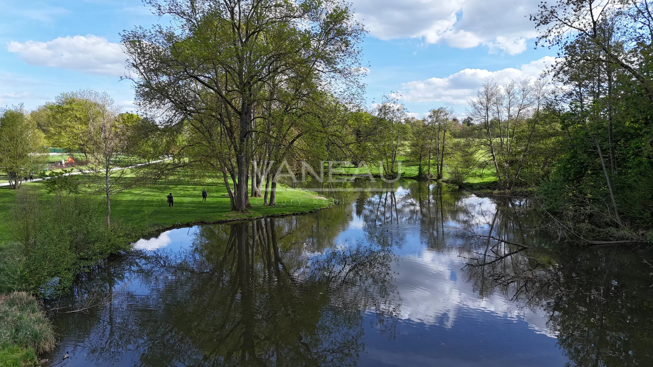 Scenic park by a calm river with tall trees and reflections on the water; a few people walk on the grassy bank under a blue sky.