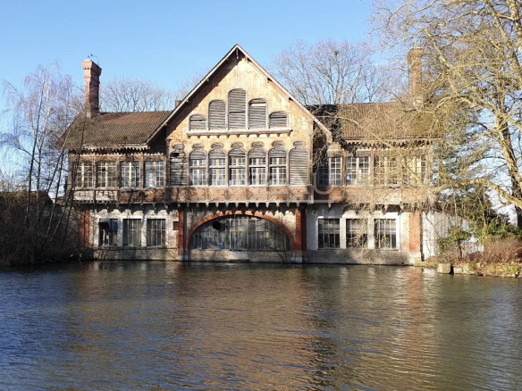 Old brick riverside building with a gabled roof and many arched windows, mirrored in calm water with leafless trees and a clear blue sky behind it.