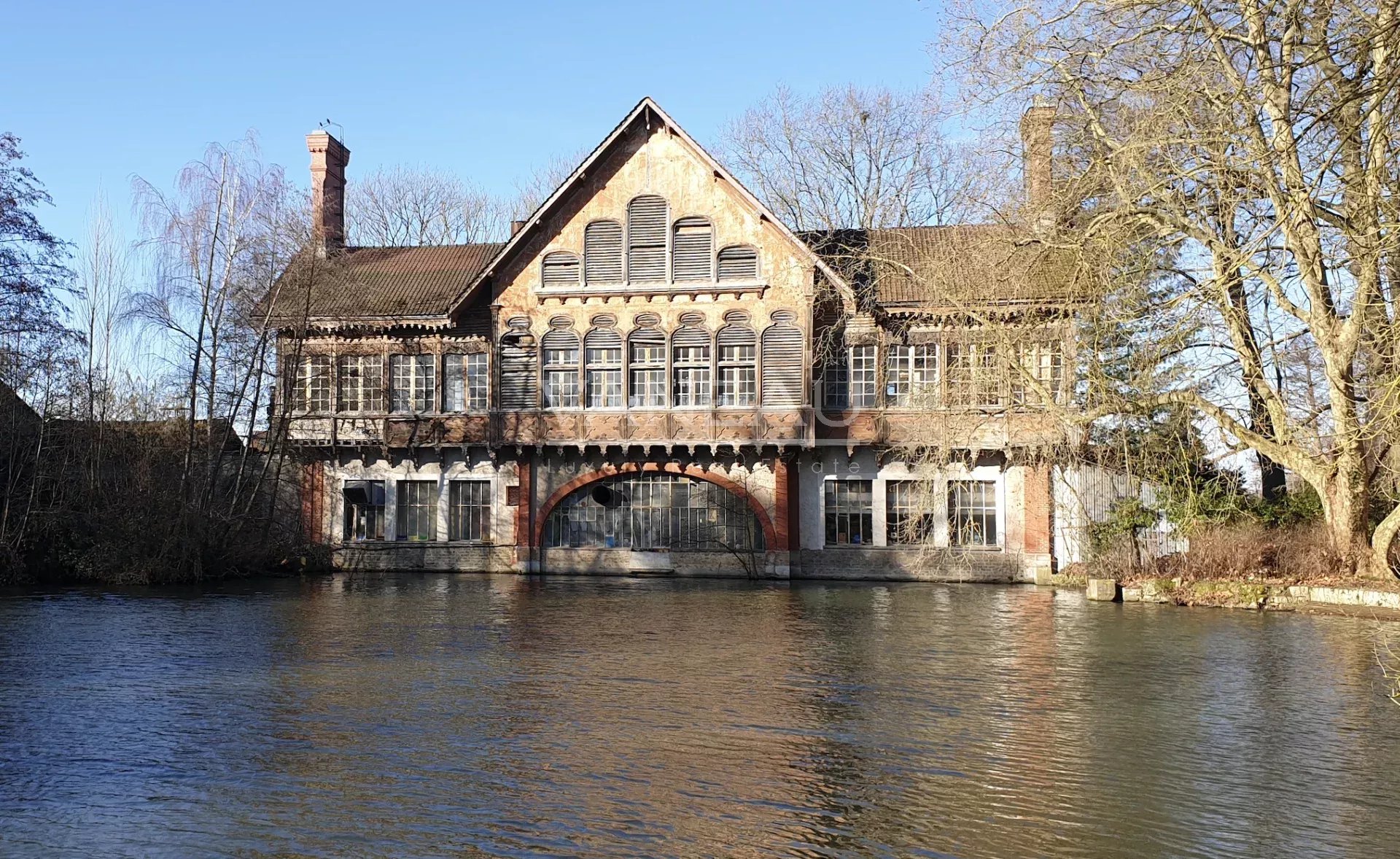 f05b35347faaa50b9348d0ae06561a16_bc4a251304_1920-original Old brick riverside building with a gabled roof and many arched windows, mirrored in calm water with leafless trees and a clear blue sky behind it.