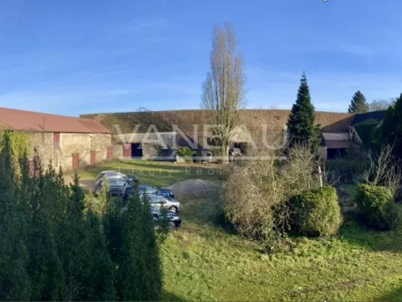 Wide view of a rural estate with red-roofed barns, a gravel driveway, and several parked cars surrounded by tall evergreens under a clear blue sky.