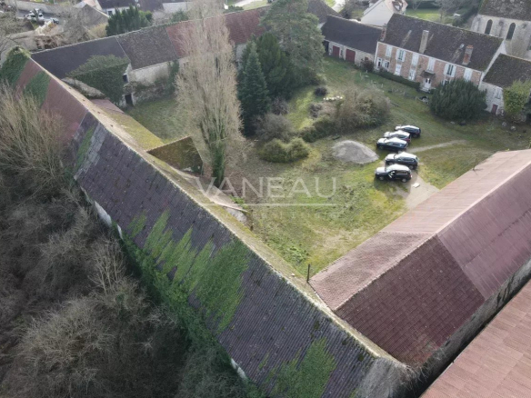 Aerial view of a historic village with steep, slate roofs, leafless trees, and a grassy yard where several cars are parked near stone buildings.