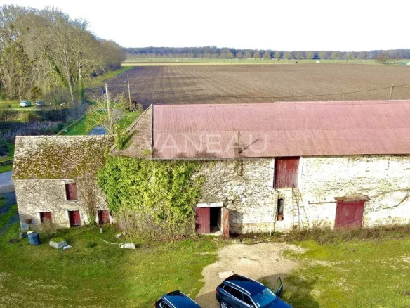 Aerial view of a weathered stone barn with red doors and ivy growing up the wall, in a rural farmland setting with fields beyond.