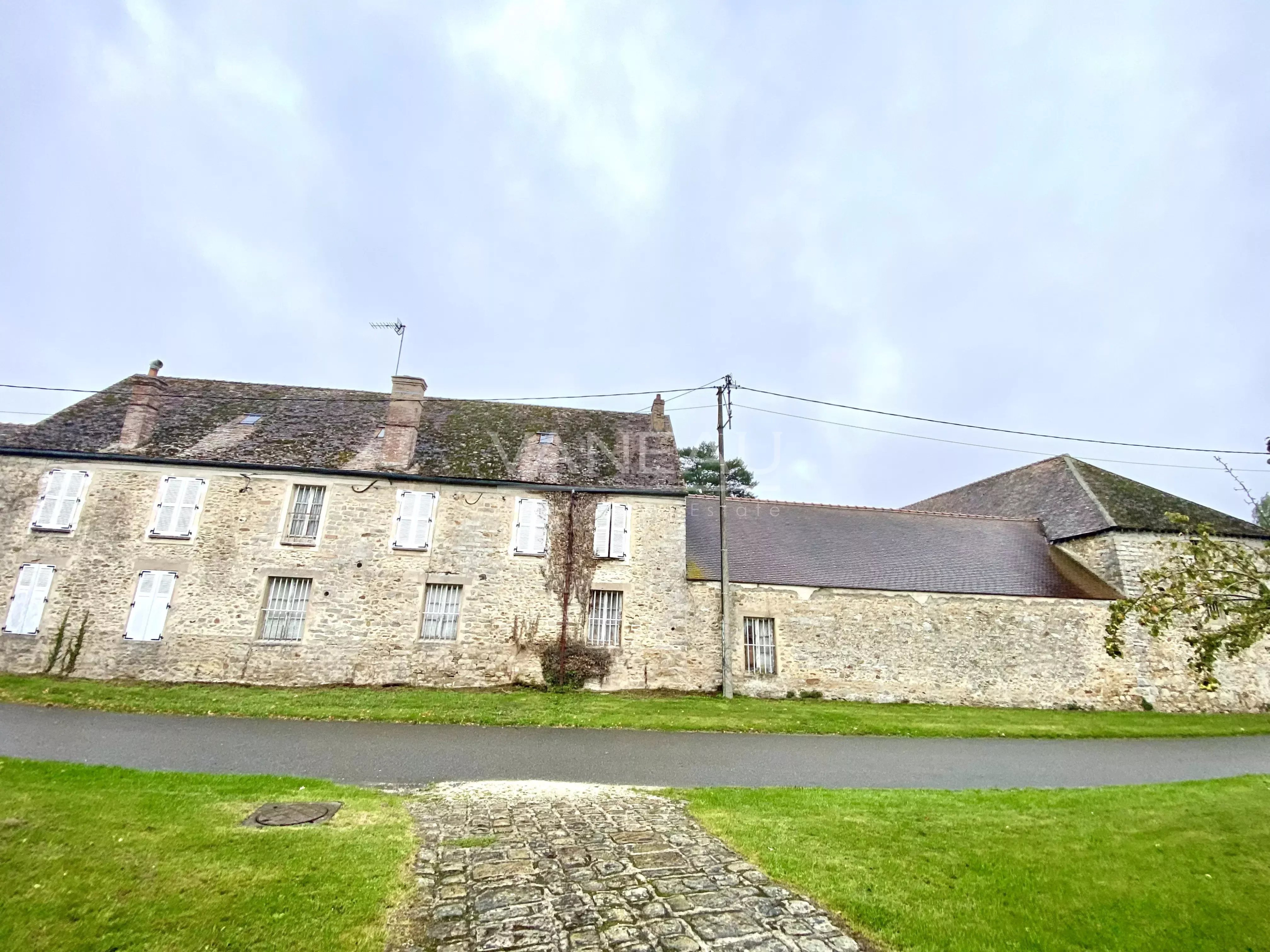 f6a593af7b09cd77c1759584b27a8170_d9921c8d25_4032-original Stone building with rows of small windows and white shutters, mossy roof, set along a grassy yard and cobblestone path under a cloudy sky.