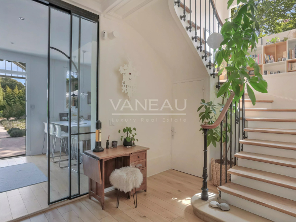 Bright foyer with a wooden desk, fluffy stool, and a large indoor plant next to a curved staircase and glass doors to the outside garden view