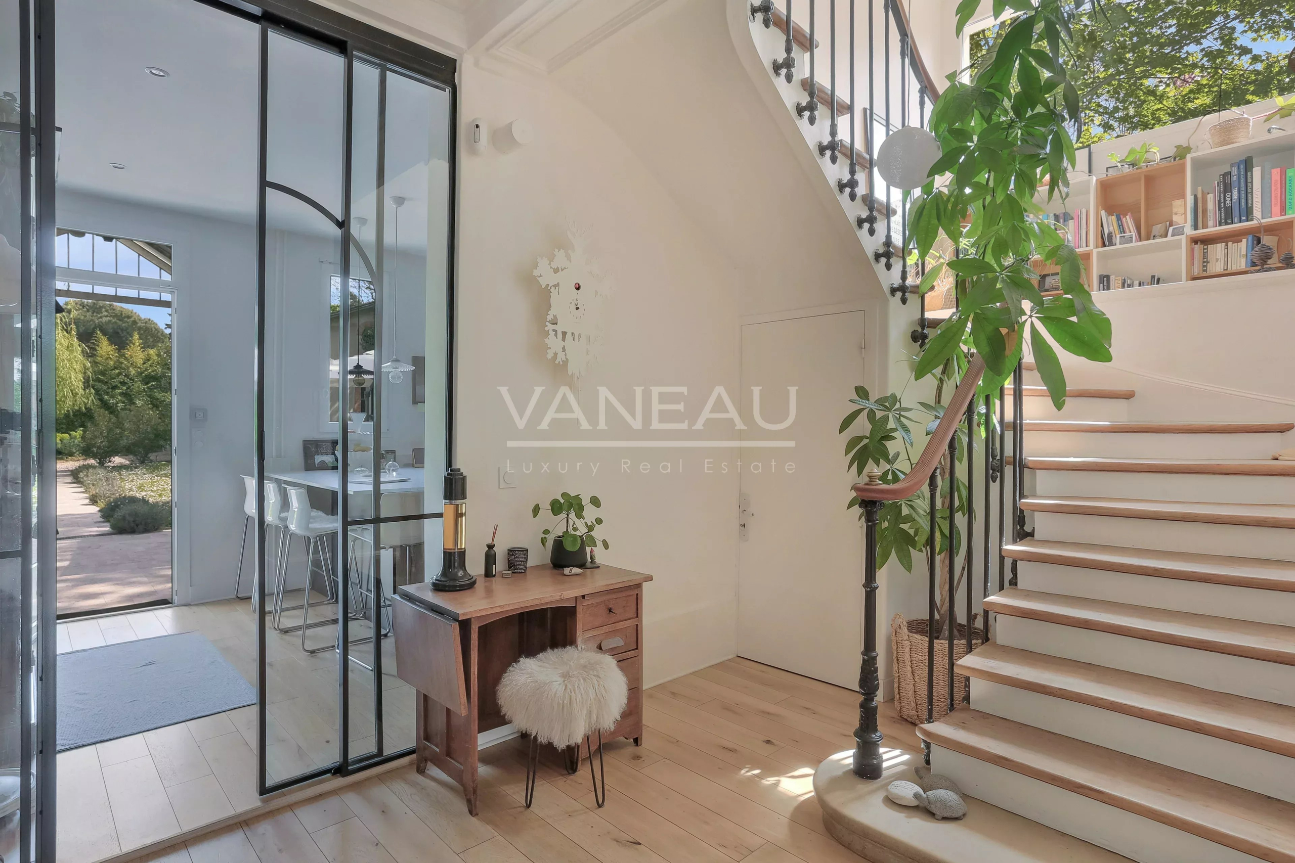 Bright foyer with a wooden desk, fluffy stool, and a large indoor plant next to a curved staircase and glass doors to the outside garden view