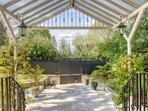 Covered patio with a glass roof, white wooden supports, and hanging lanterns, opening to a garden with potted plants and a black iron gate.