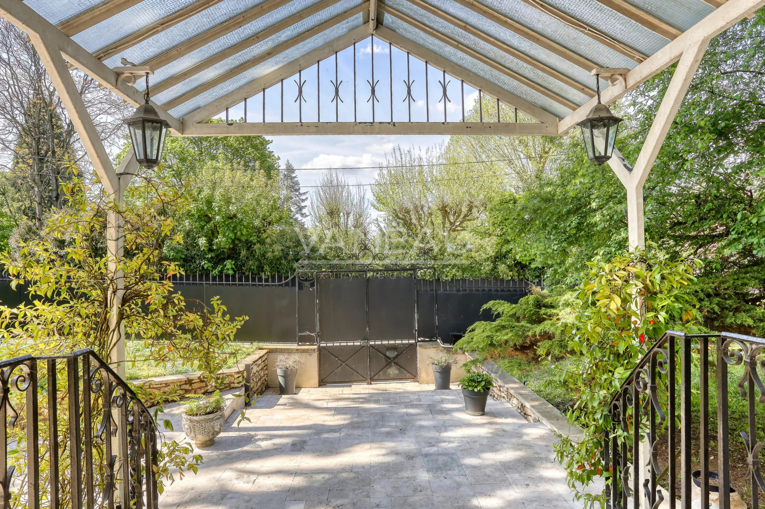 Covered patio with a glass roof, white wooden supports, and hanging lanterns, opening to a garden with potted plants and a black iron gate.