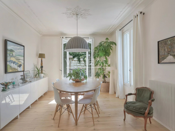 Bright dining room with a round wooden table, white chairs, and a large hanging pendant light, flanked by plants near open doors.