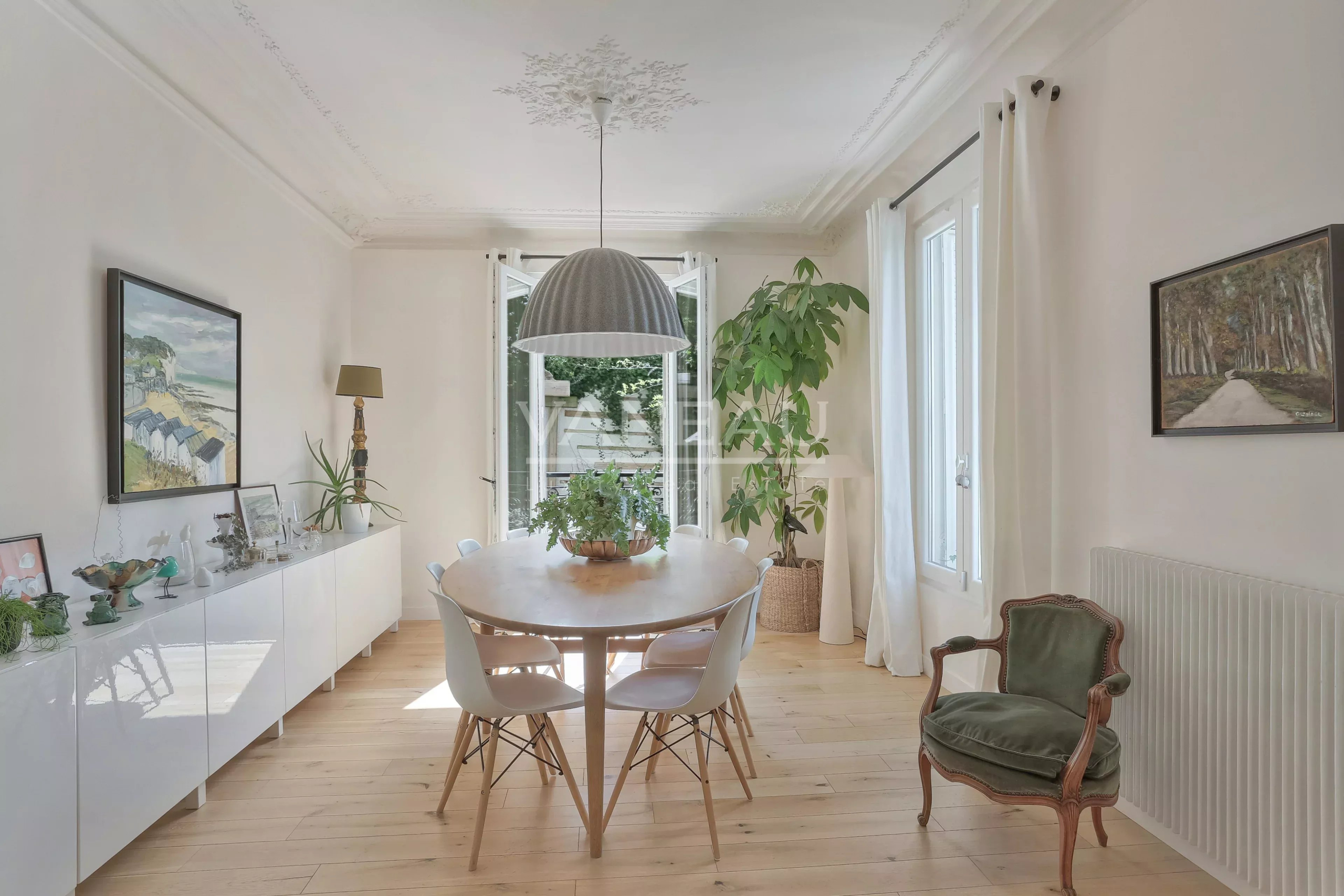 Bright dining room with a round wooden table, white chairs, and a large hanging pendant light, flanked by plants near open doors.