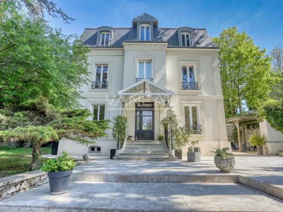 Elegant light-gray mansion with a central entrance, stone steps, and a manicured garden beneath a clear blue sky.
