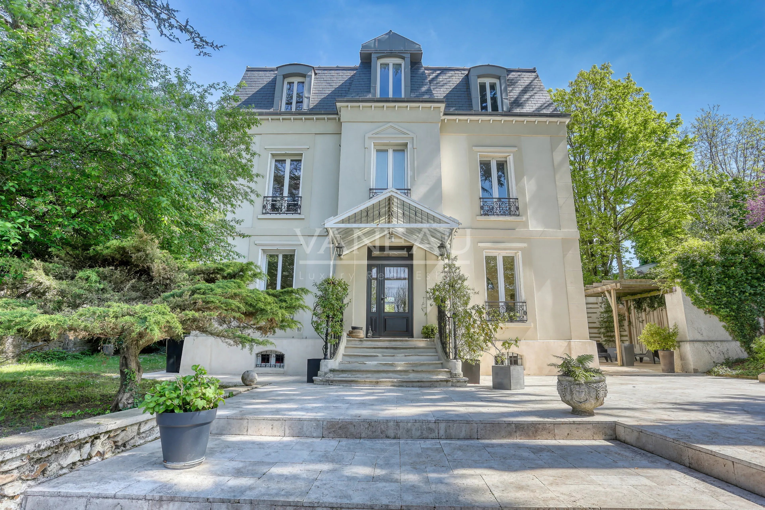 Elegant light-gray mansion with a central entrance, stone steps, and a manicured garden beneath a clear blue sky.