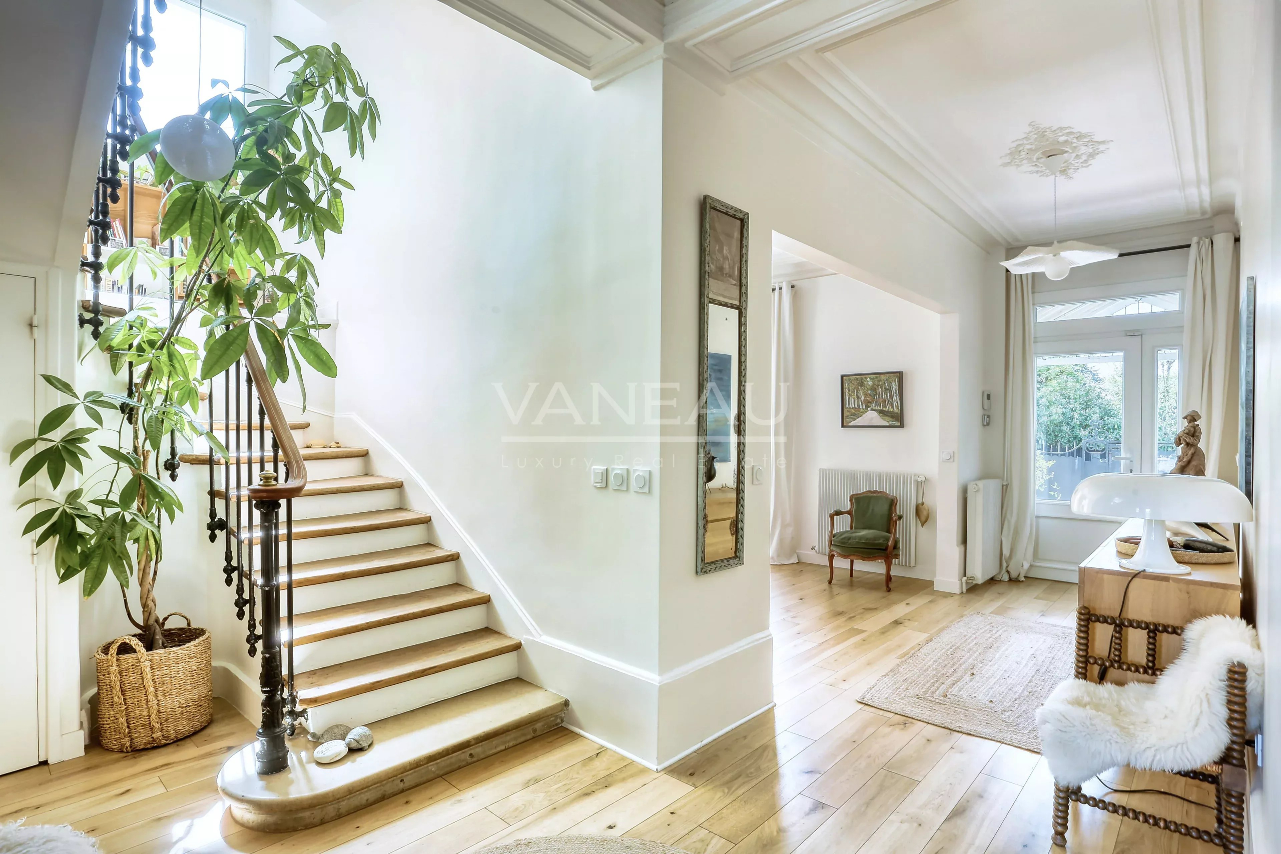Bright foyer with curved stairway on the left, a tall potted plant, and wicker basket by the wall.