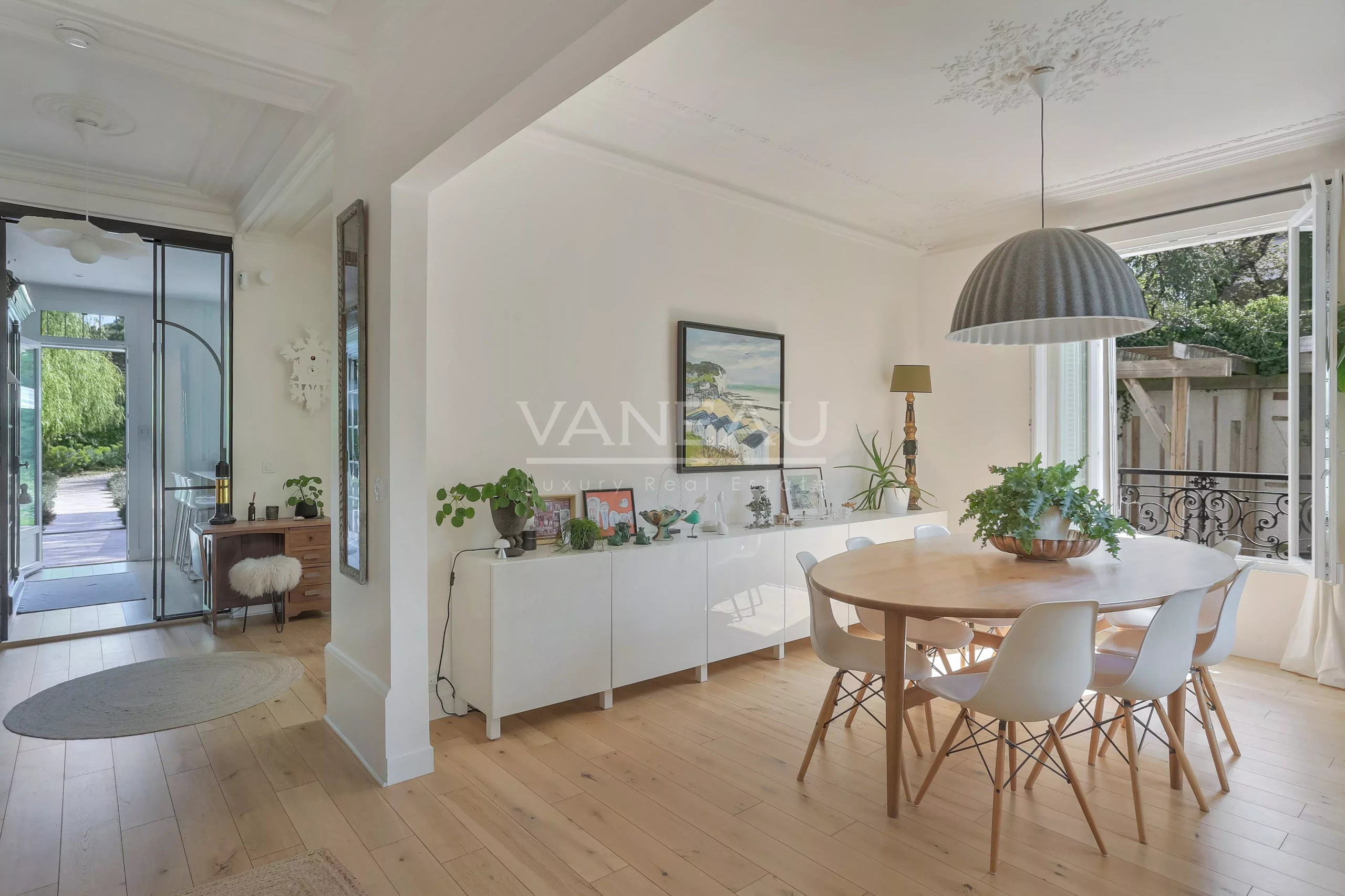 Bright dining area with a round wooden table, white chairs, and open glass doors to a balcony with greenery outside.
