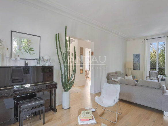 Bright living room with a glossy black upright piano, tall cactus in a white planter, and beige sofas under natural light from a large window with white curtains.
