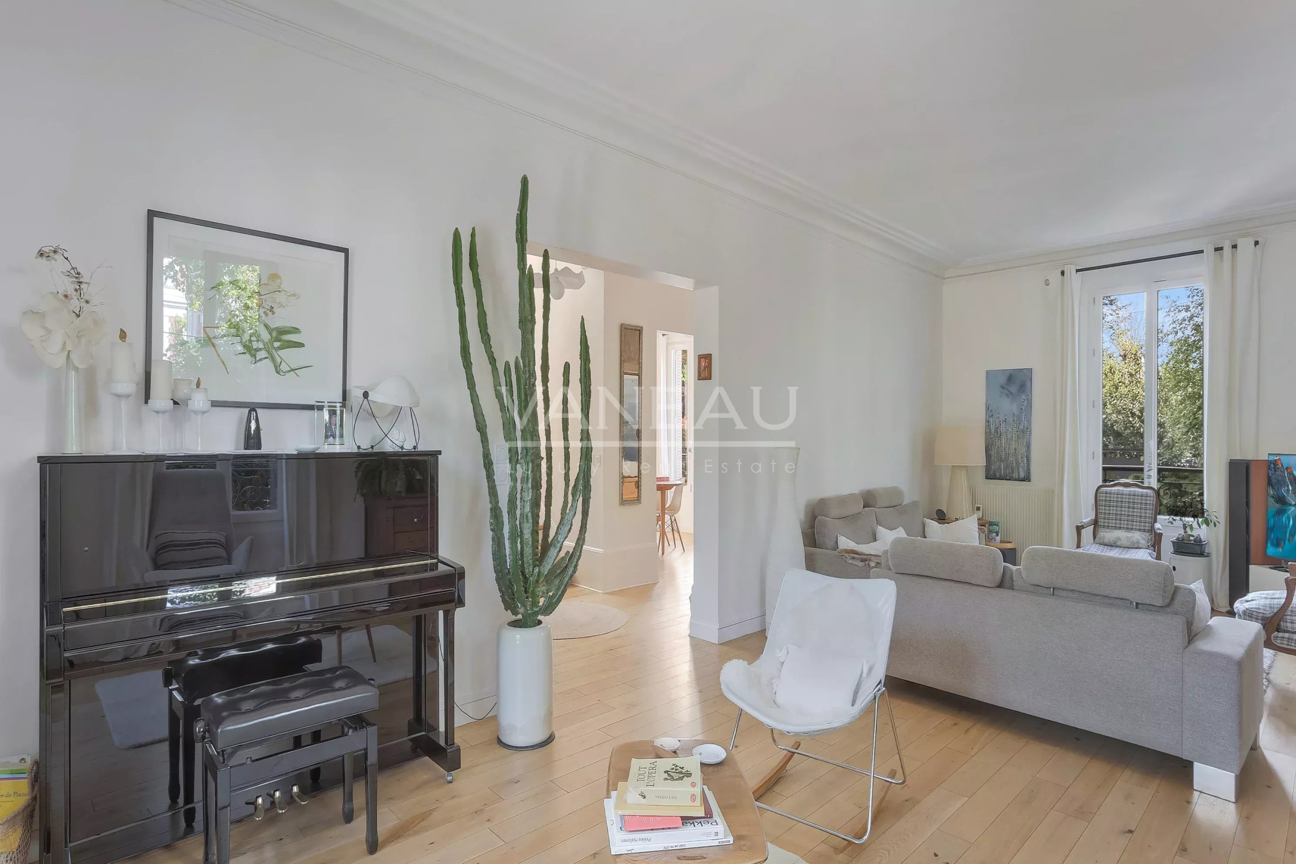 Bright living room with a glossy black upright piano, tall cactus in a white planter, and beige sofas under natural light from a large window with white curtains.