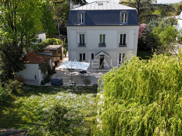 Aerial view of a white three-story house with a blue mansard roof, surrounded by lush trees and a sunny garden courtyard with a white canopy and outdoor seating.