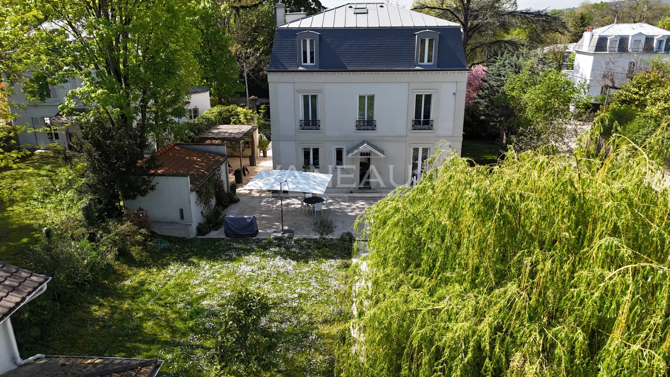 Aerial view of a white three-story house with a blue mansard roof, surrounded by lush trees and a sunny garden courtyard with a white canopy and outdoor seating.