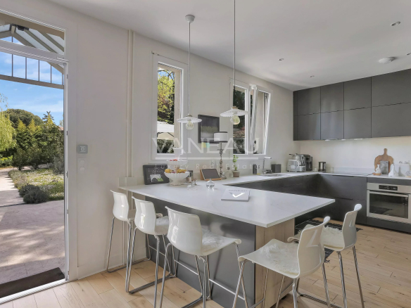 Modern open kitchen with a white island, gray cabinets, and white bar stools facing outdoors through a glass door.
