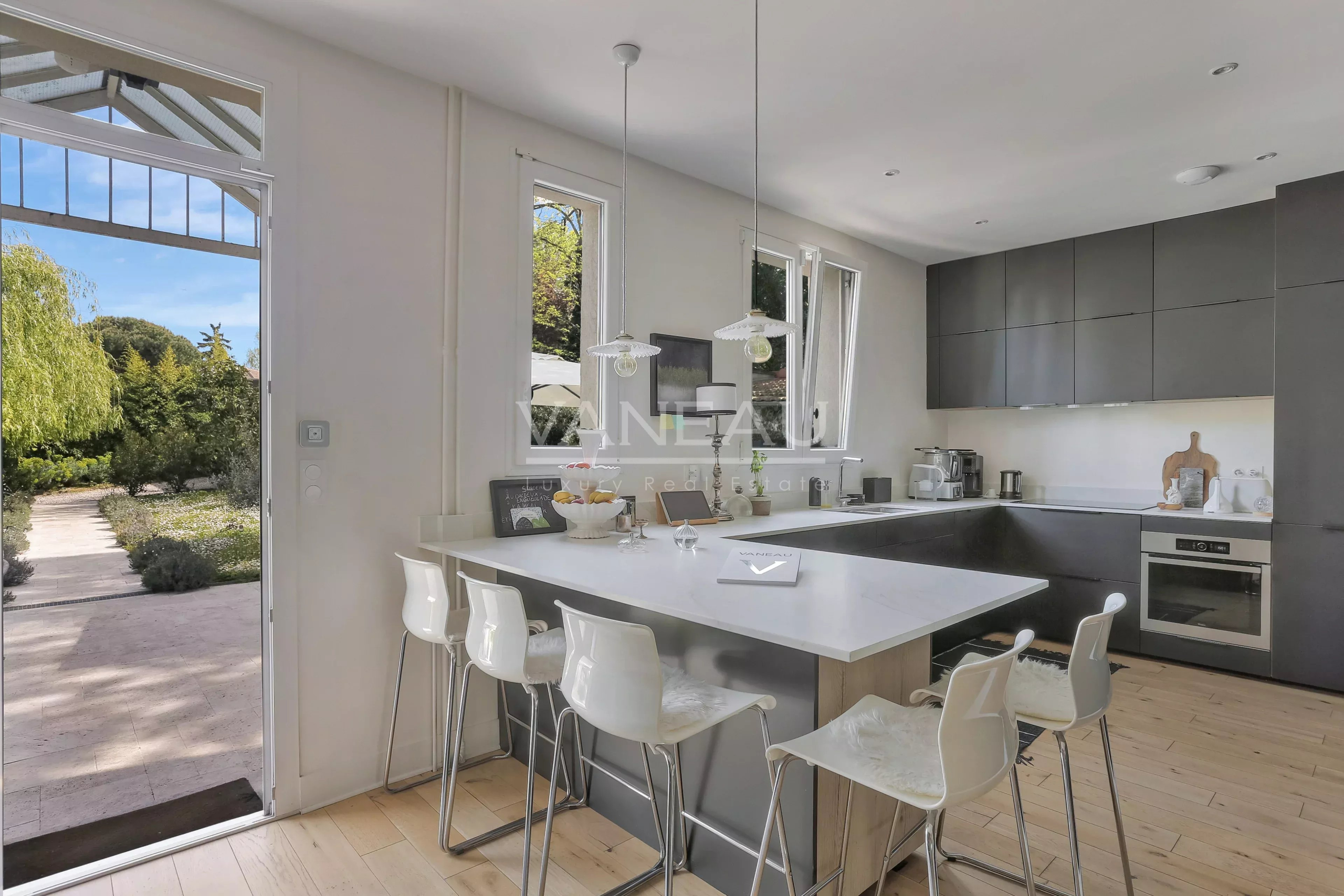 Modern open kitchen with a white island, gray cabinets, and white bar stools facing outdoors through a glass door.