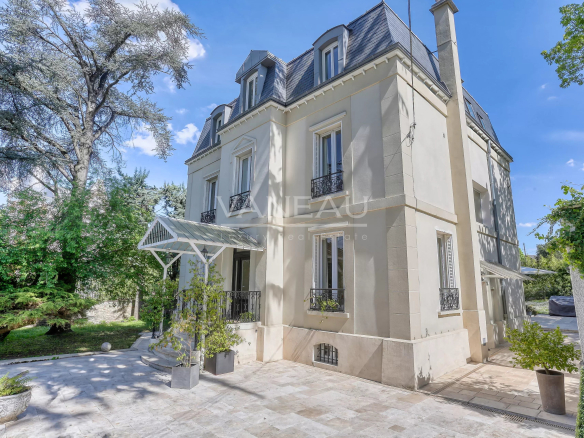 Cream-colored, three-story villa with a mansard roof and a glass canopy over the entrance, set in a landscaped courtyard.