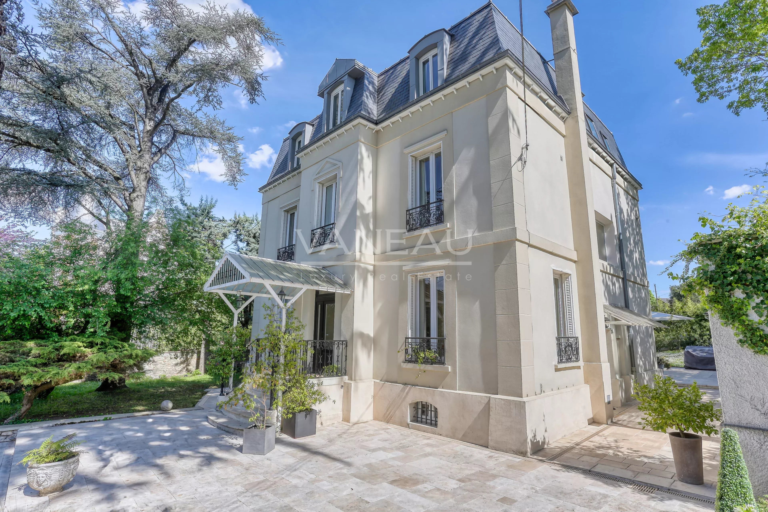 Cream-colored, three-story villa with a mansard roof and a glass canopy over the entrance, set in a landscaped courtyard.