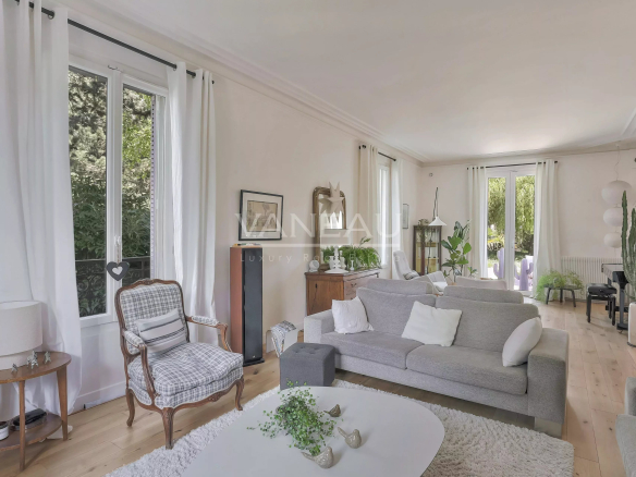 Bright, airy living room with a light gray sofa, white curtains, and large windows letting in greenery outside.