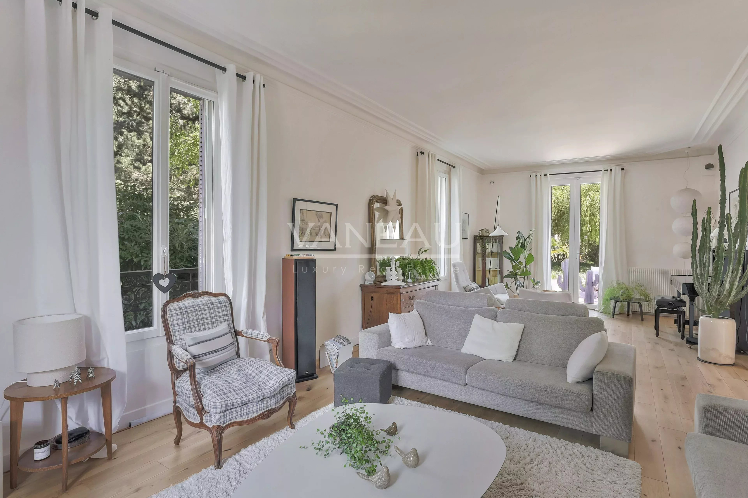 Bright, airy living room with a light gray sofa, white curtains, and large windows letting in greenery outside.