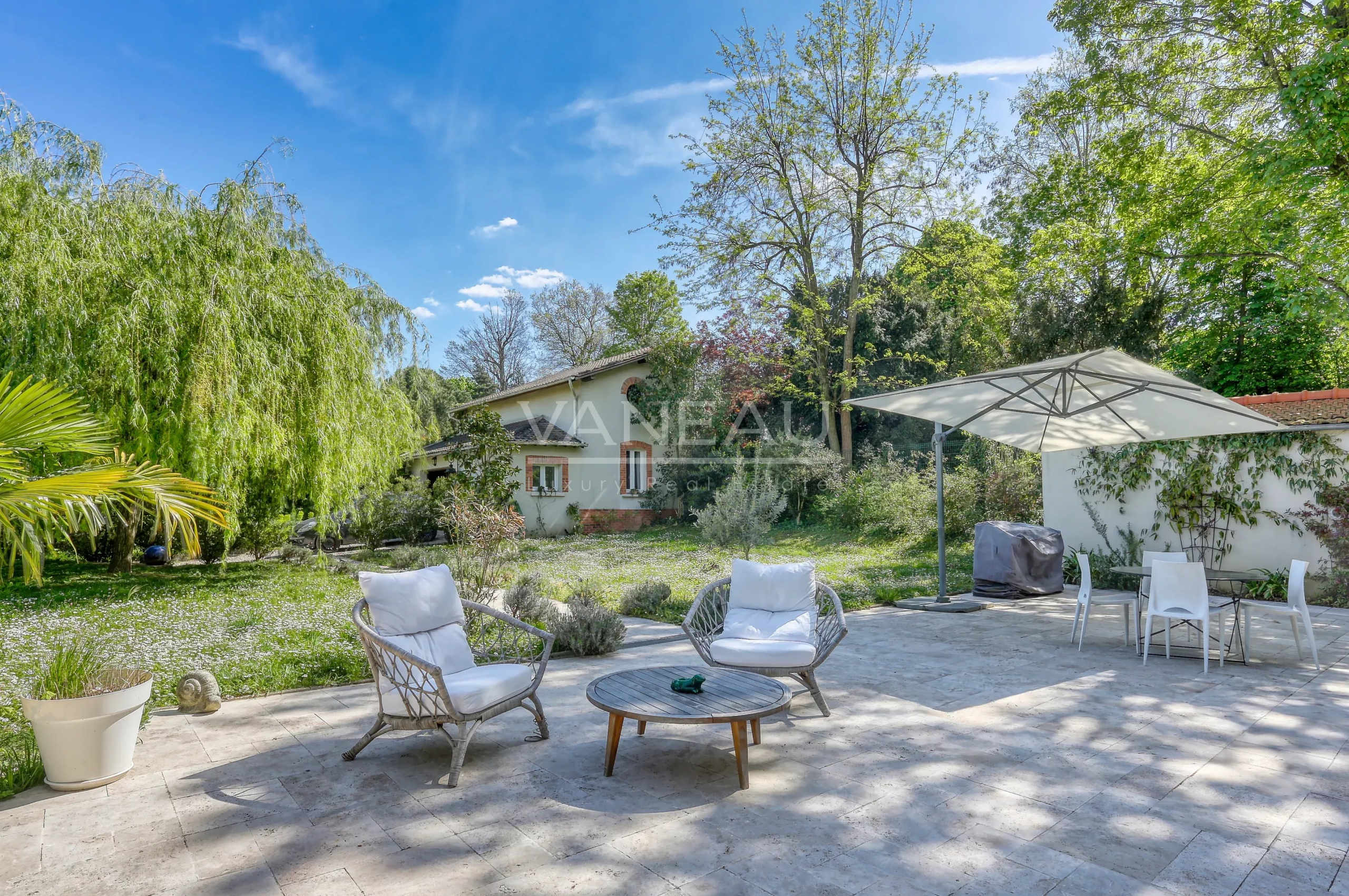 Sunny backyard patio with two wicker chairs and a round table under a large umbrella, white cushions, and a garden beyond the concrete patio.