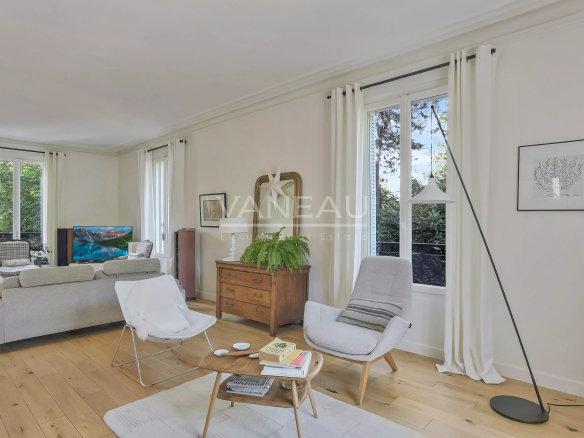 Bright living room with white walls and large windows, featuring a gray sofa and chairs around a wooden coffee table.