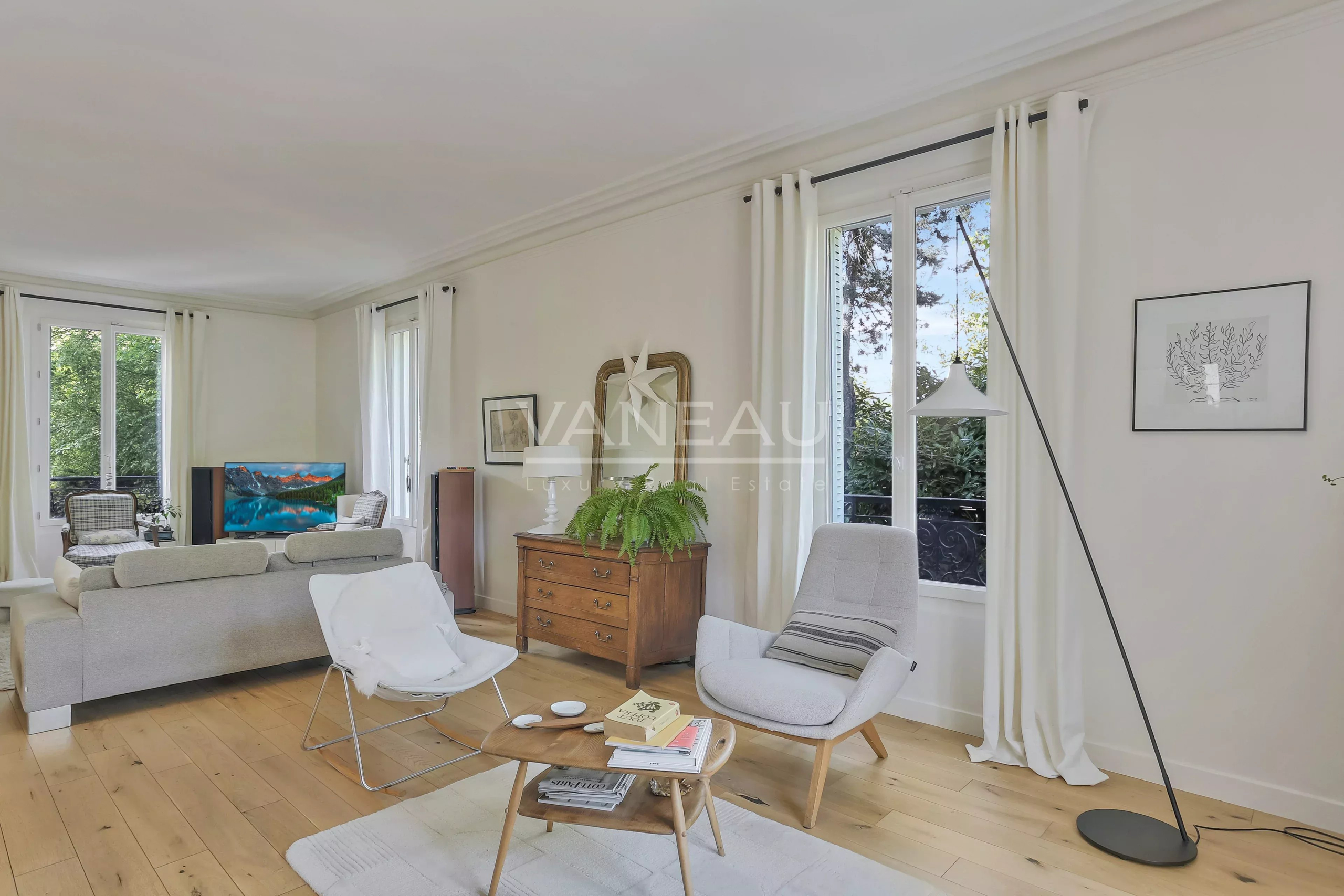 Bright living room with white walls and large windows, featuring a gray sofa and chairs around a wooden coffee table.
