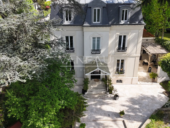 Aerial view of a beige, four-story house with a dark slate roof, centered entrance with stairs and a glass canopy, and a stone-patio courtyard surrounded by lush trees and shrubs.