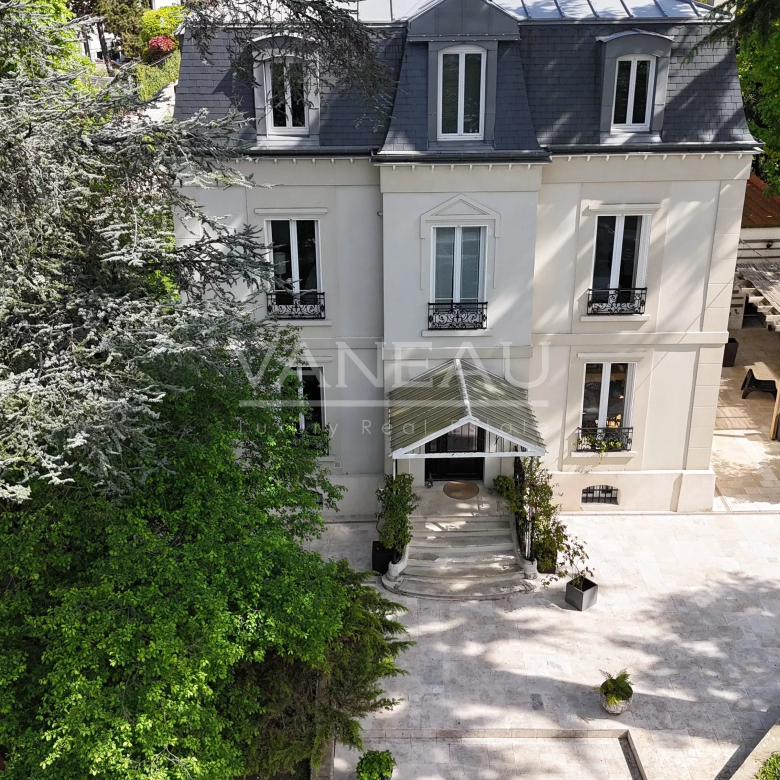 Aerial view of a beige, four-story house with a dark slate roof, centered entrance with stairs and a glass canopy, and a stone-patio courtyard surrounded by lush trees and shrubs.