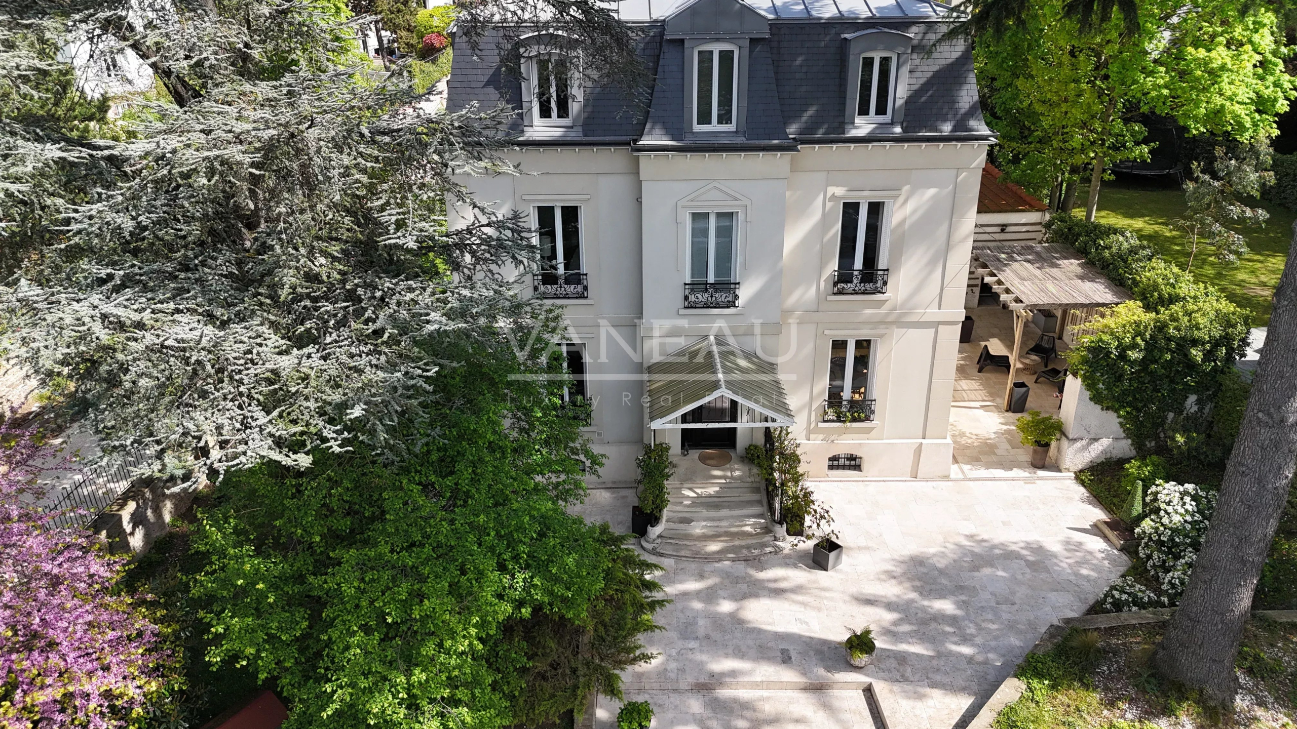 Aerial view of a beige, four-story house with a dark slate roof, centered entrance with stairs and a glass canopy, and a stone-patio courtyard surrounded by lush trees and shrubs.