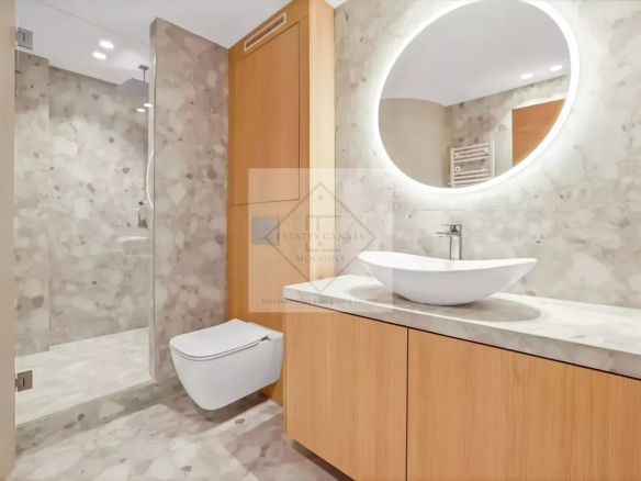 Modern bathroom featuring a round backlit mirror above a marble countertop with a white vessel sink and wooden vanity. A glass-enclosed shower and wall-mounted toilet complete the space.