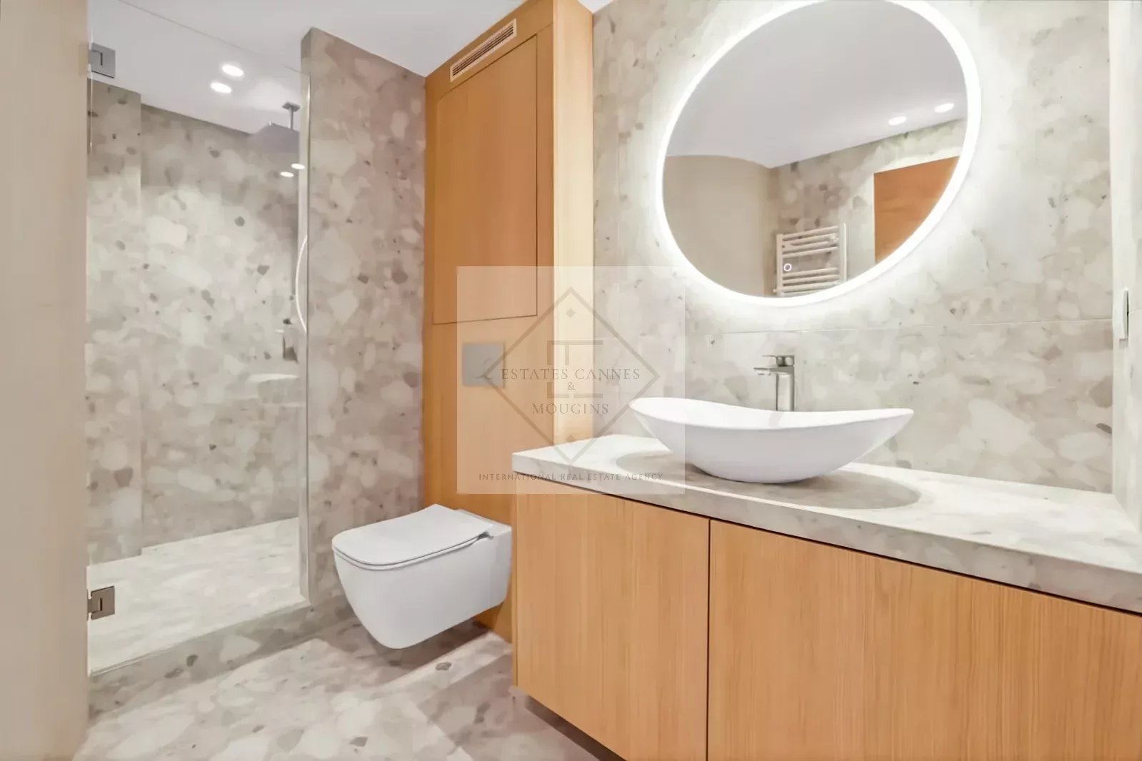 Modern bathroom featuring a round backlit mirror above a marble countertop with a white vessel sink and wooden vanity. A glass-enclosed shower and wall-mounted toilet complete the space.